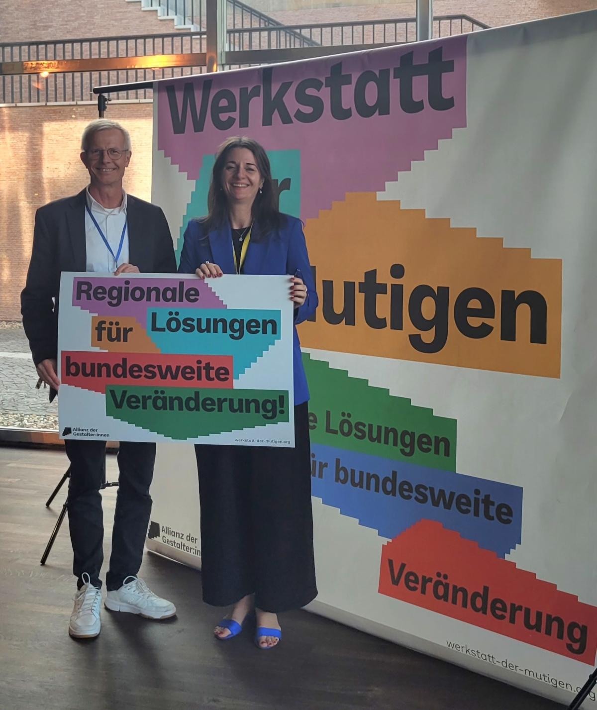Zwei Personen halten ein buntes Schild mit "Regionale Lösungen für bundesweite Veränderung"